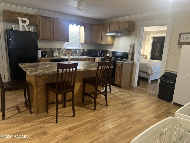 kitchen with under cabinet range hood, a kitchen breakfast bar, light wood-style floors, black appliances, and crown molding