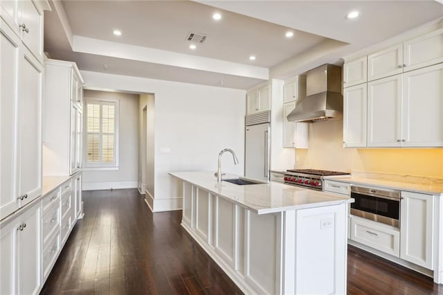 kitchen with built in fridge, a kitchen island with sink, a sink, wall chimney exhaust hood, and cooktop