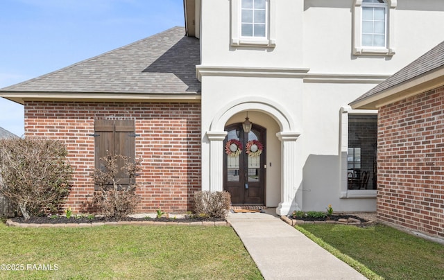 view of exterior entry featuring a yard, brick siding, roof with shingles, and stucco siding