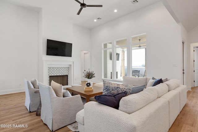 living room with baseboards, a tile fireplace, a towering ceiling, ceiling fan, and light wood-style flooring