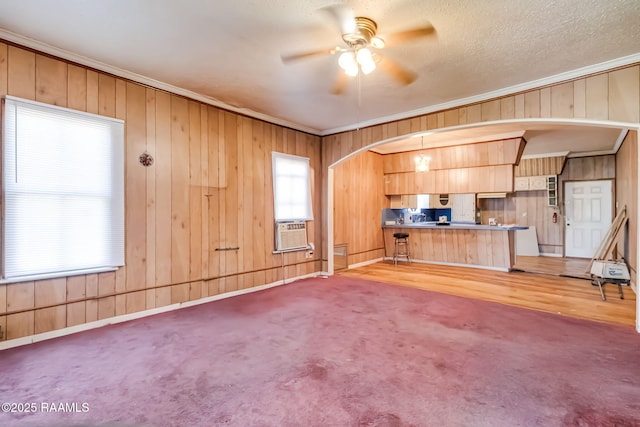 unfurnished living room featuring wooden walls, arched walkways, a ceiling fan, ornamental molding, and cooling unit