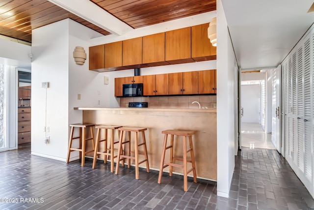 kitchen featuring black microwave, a breakfast bar area, brown cabinetry, tasteful backsplash, and beamed ceiling