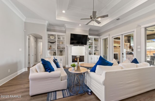 living room with arched walkways, a tray ceiling, wood finished floors, a lit fireplace, and baseboards