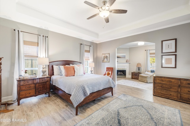 bedroom with crown molding, light wood-type flooring, a tray ceiling, a fireplace, and arched walkways