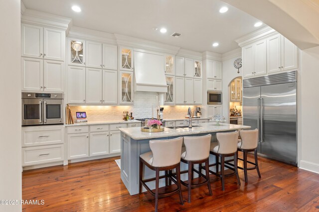 kitchen with arched walkways, white cabinets, custom range hood, and built in appliances