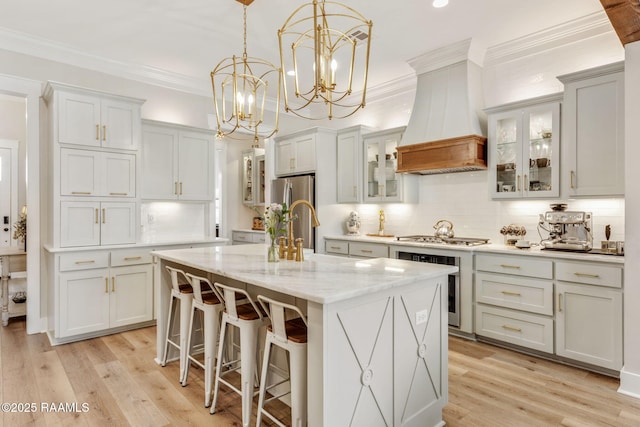 kitchen featuring premium range hood, light wood-type flooring, a kitchen island with sink, ornamental molding, and appliances with stainless steel finishes