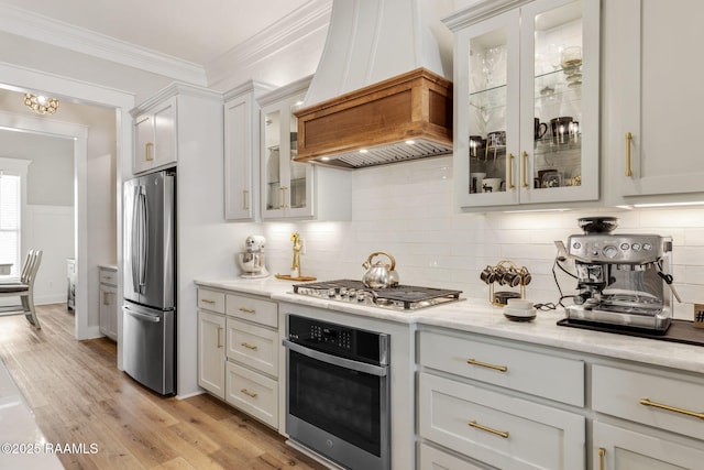 kitchen featuring light wood-type flooring, custom range hood, backsplash, stainless steel appliances, and crown molding