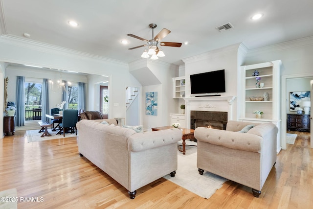 living area featuring visible vents, stairway, light wood-type flooring, a lit fireplace, and ceiling fan with notable chandelier