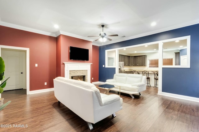 living area featuring wood finished floors, baseboards, a warm lit fireplace, ceiling fan, and ornamental molding