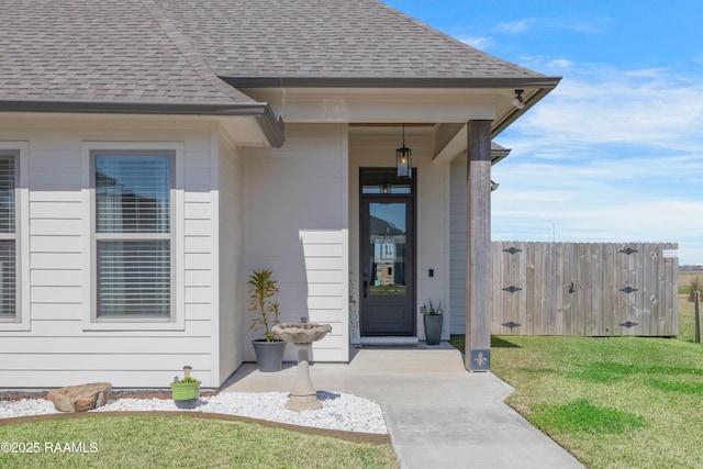 view of exterior entry with a lawn, roof with shingles, and fence