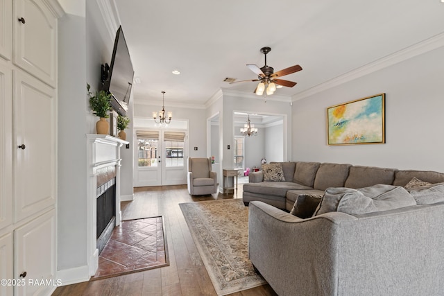 living room featuring a tiled fireplace, dark wood-style flooring, crown molding, and ceiling fan with notable chandelier