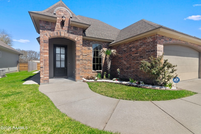 french country style house with fence, a shingled roof, a front lawn, a garage, and brick siding