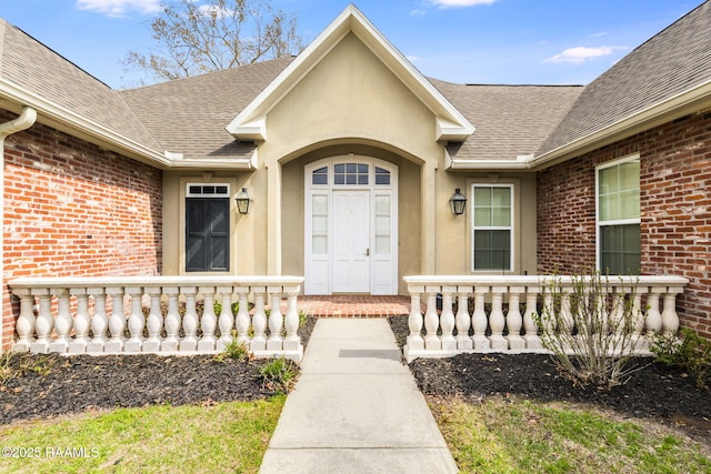 view of exterior entry with stucco siding, brick siding, a porch, and a shingled roof