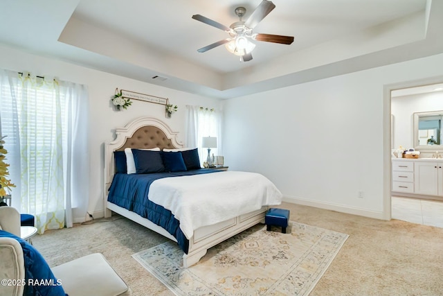 bedroom with baseboards, a raised ceiling, light colored carpet, and ensuite bathroom