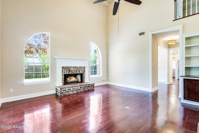 unfurnished living room featuring visible vents, a ceiling fan, wood finished floors, a high ceiling, and a brick fireplace
