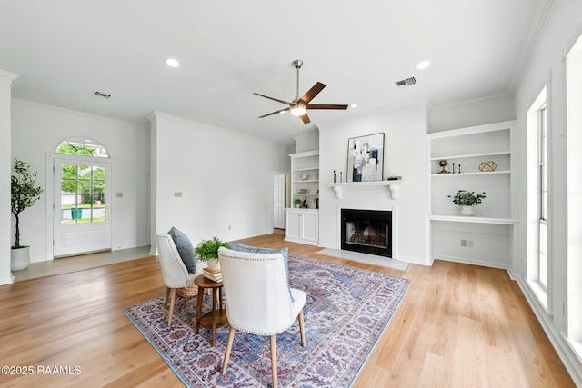 living area featuring a ceiling fan, visible vents, a fireplace with flush hearth, light wood-style floors, and ornamental molding