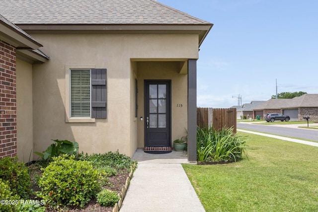 entrance to property featuring stucco siding, roof with shingles, fence, and a yard