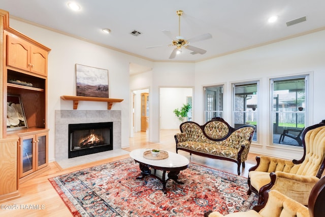living room featuring a tiled fireplace, light wood-style flooring, ornamental molding, ceiling fan, and visible vents