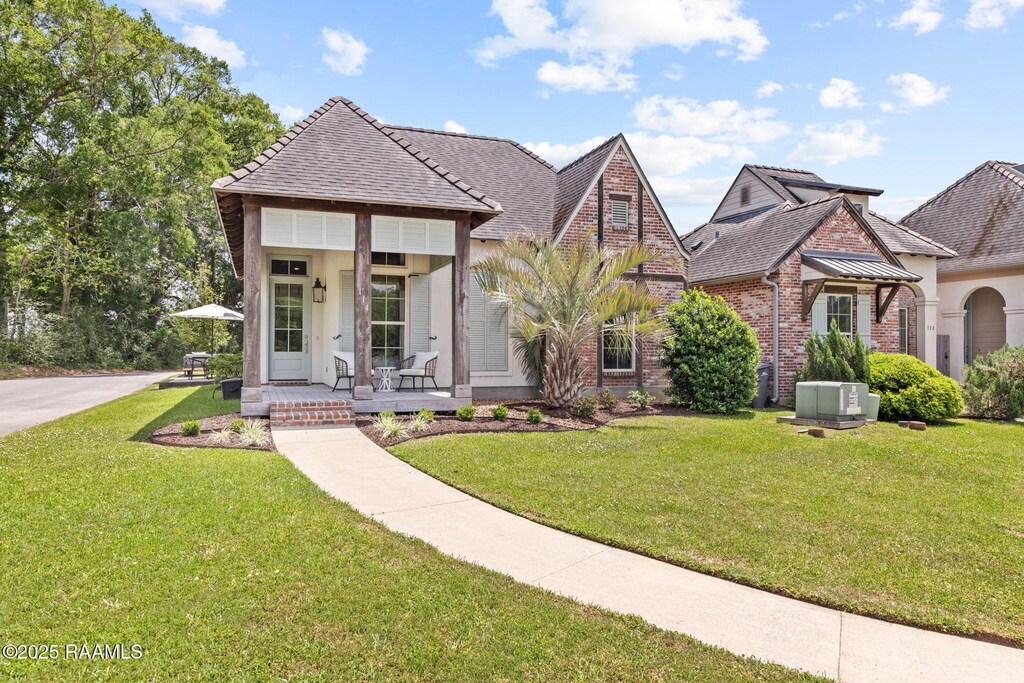 french country home featuring roof with shingles, covered porch, central AC unit, brick siding, and a front lawn