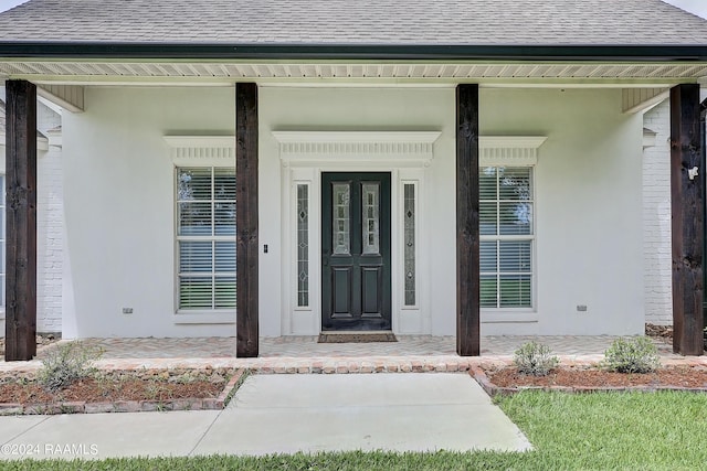 doorway to property with a porch, brick siding, stucco siding, and roof with shingles
