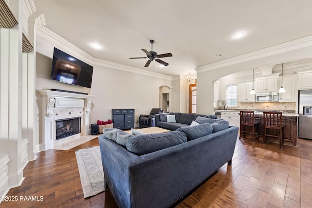 living area featuring ceiling fan, arched walkways, a tile fireplace, dark wood-style floors, and ornamental molding