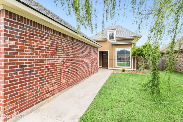 property entrance featuring a lawn, fence, roof with shingles, and brick siding