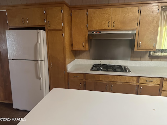 kitchen with brown cabinetry, freestanding refrigerator, gas cooktop, light countertops, and under cabinet range hood
