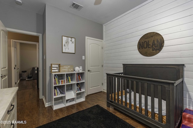 bedroom with dark wood-style floors, ceiling fan, visible vents, wood walls, and a crib