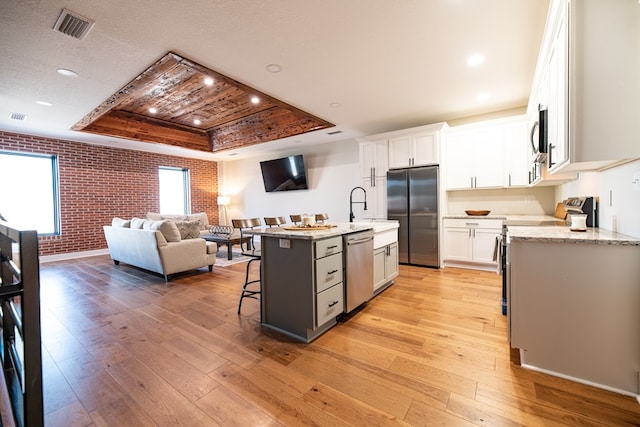 kitchen featuring appliances with stainless steel finishes, brick wall, a tray ceiling, a kitchen island with sink, and white cabinets