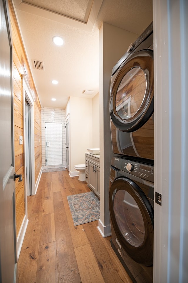 washroom featuring a textured ceiling, wooden walls, light hardwood / wood-style flooring, and stacked washer / drying machine