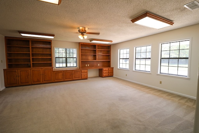 unfurnished living room featuring light carpet, built in desk, plenty of natural light, and ceiling fan