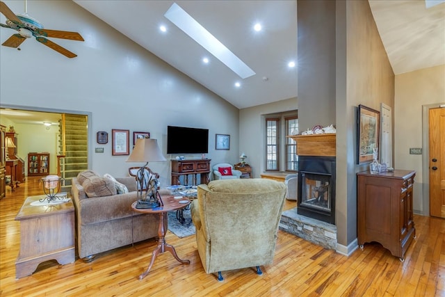 living area featuring high vaulted ceiling, a skylight, stairs, light wood finished floors, and a glass covered fireplace