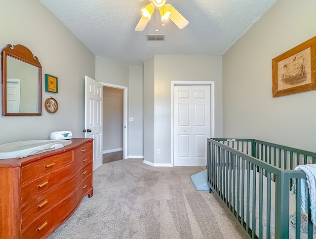 bedroom with light colored carpet, a nursery area, a closet, ceiling fan, and a textured ceiling