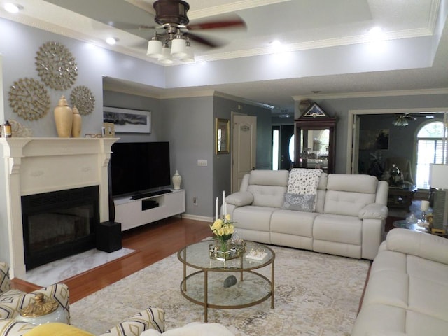 living area with ornamental molding, a fireplace with flush hearth, wood finished floors, a raised ceiling, and a ceiling fan
