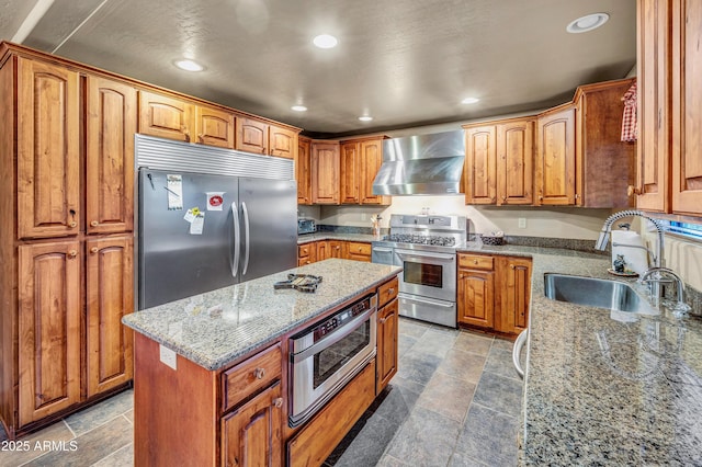 kitchen with stainless steel appliances, stone finish flooring, a sink, wall chimney range hood, and light stone countertops