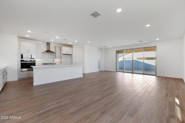 kitchen featuring oven, tasteful backsplash, a kitchen island with sink, white cabinets, and wall chimney exhaust hood