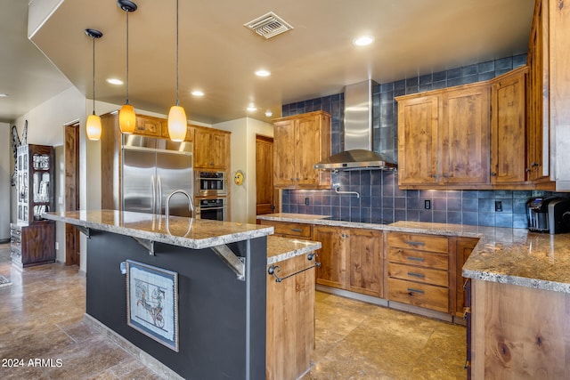 kitchen with appliances with stainless steel finishes, a breakfast bar, pendant lighting, a center island with sink, and wall chimney range hood