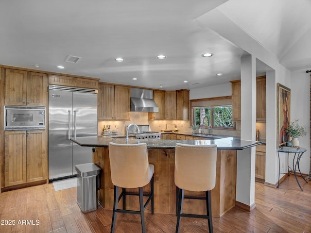 kitchen with brown cabinets, a sink, built in appliances, light wood finished floors, and wall chimney exhaust hood
