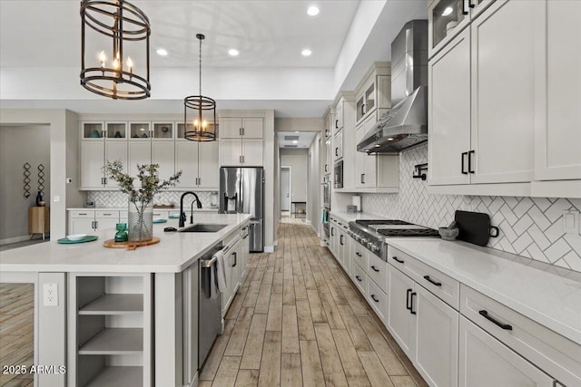 kitchen with sink, hanging light fixtures, appliances with stainless steel finishes, a kitchen island with sink, and wall chimney range hood