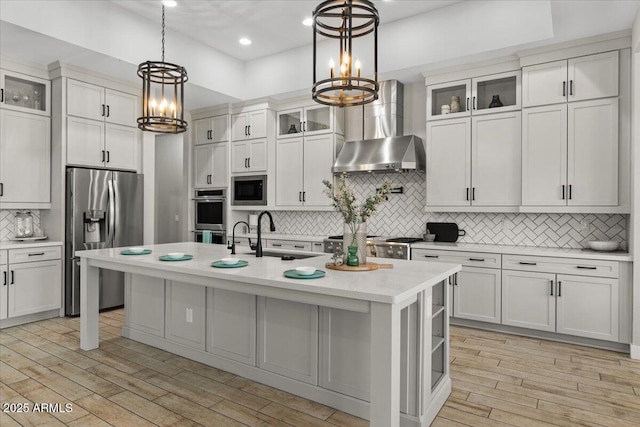 kitchen with decorative light fixtures, an island with sink, white cabinets, stainless steel appliances, and wall chimney exhaust hood