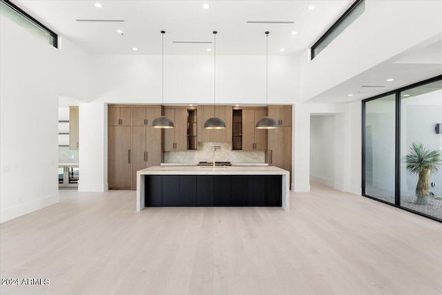 kitchen featuring light hardwood / wood-style flooring, tasteful backsplash, decorative light fixtures, a large island with sink, and a towering ceiling