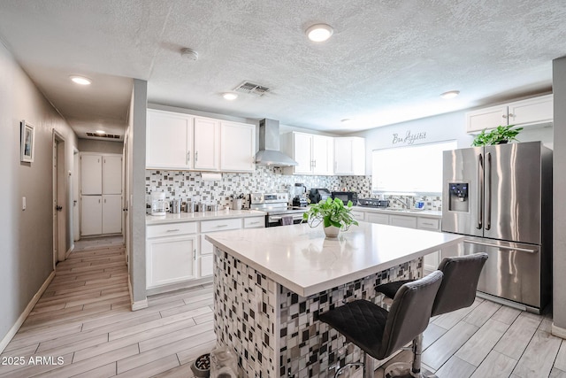 kitchen featuring a kitchen bar, stainless steel appliances, wall chimney range hood, white cabinets, and a center island