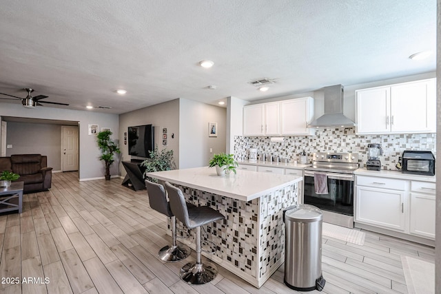 kitchen featuring stainless steel electric range, white cabinets, wall chimney range hood, and a kitchen island