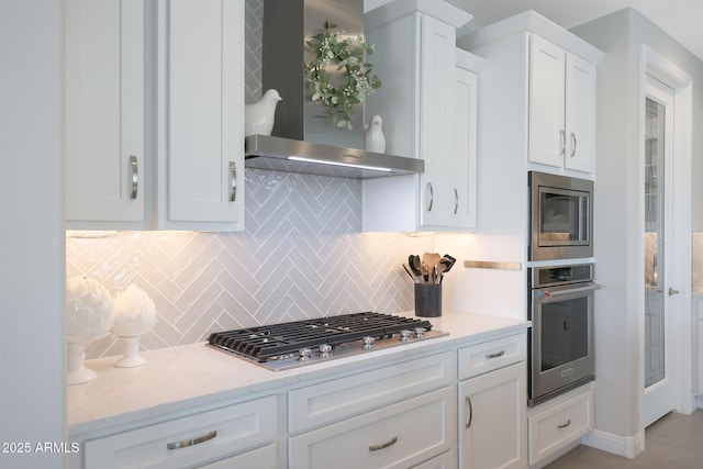 kitchen featuring light stone counters, stainless steel appliances, white cabinetry, wall chimney range hood, and tasteful backsplash