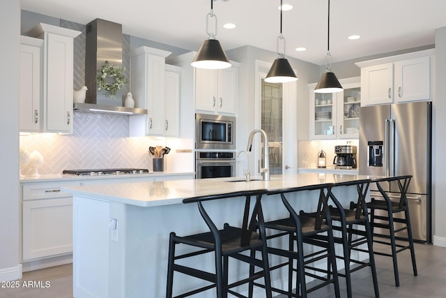 kitchen featuring stainless steel appliances, white cabinetry, light countertops, wall chimney range hood, and an island with sink