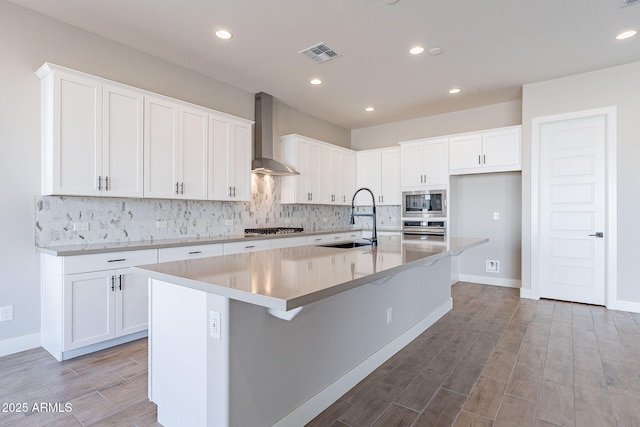 kitchen with wall chimney exhaust hood, sink, white cabinetry, and an island with sink