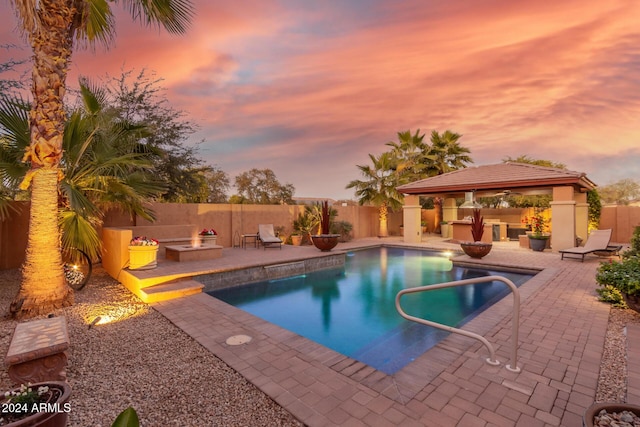 pool at dusk with an outdoor kitchen, a gazebo, a patio, and a fire pit