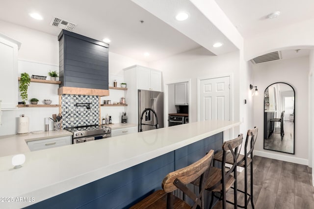 kitchen featuring visible vents, open shelves, a kitchen bar, wall chimney exhaust hood, and appliances with stainless steel finishes