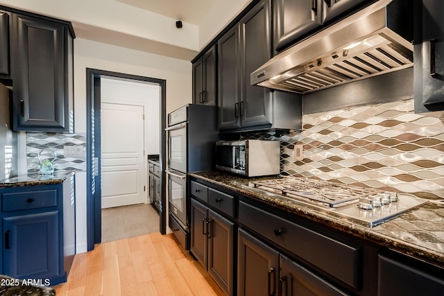 kitchen featuring appliances with stainless steel finishes, dark stone countertops, backsplash, ventilation hood, and light wood-type flooring