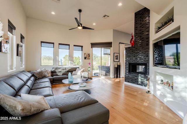 living room with ceiling fan, a high ceiling, a stone fireplace, and light wood-type flooring